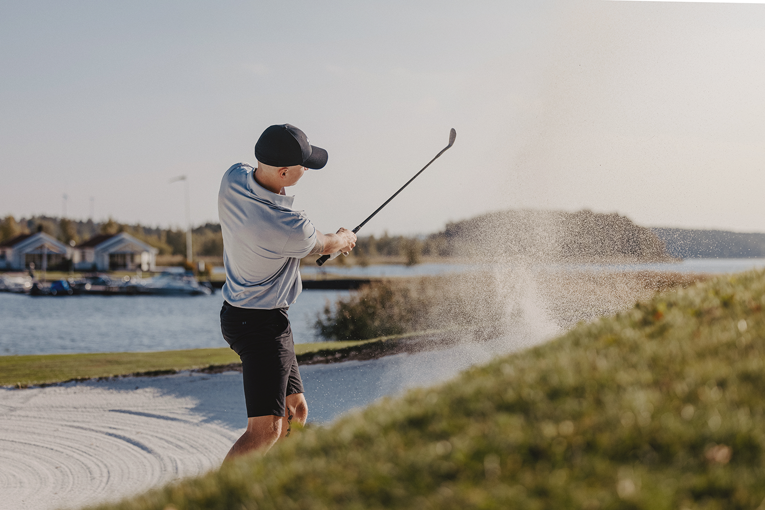 Man slår en golfboll ur en sandbunker, sanden yr och en sjö samt hus syns i bakgrunden.