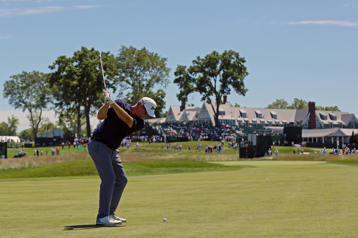 Golfspelare svingar en klubba på fairway, med publik och klubbhus i bakgrunden under en solig dag.