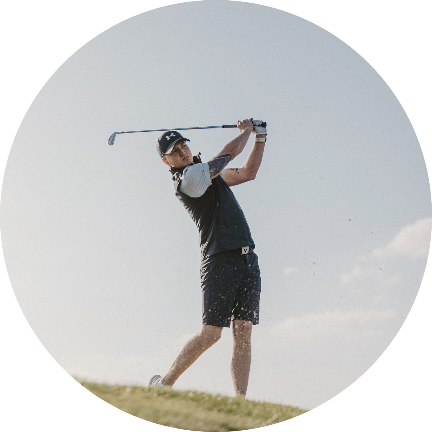 Man slår ett golfslag på en golfbana, klädd i sportkläder och keps under en klar himmel.