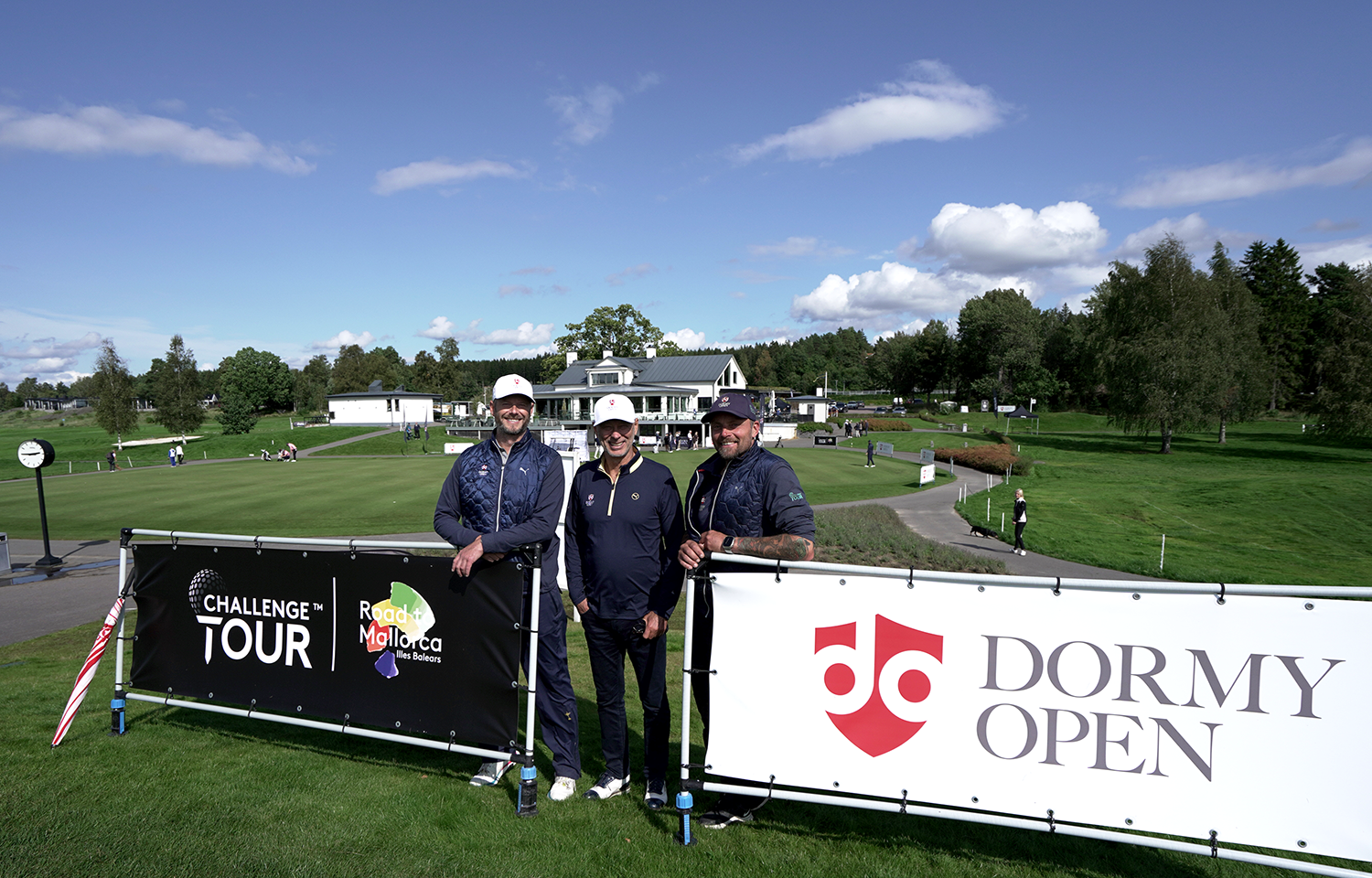 Tre män poserar framför skyltar med texten "Challenge Tour" och "Dormy Open" på en golfbana under klarblå himmel.