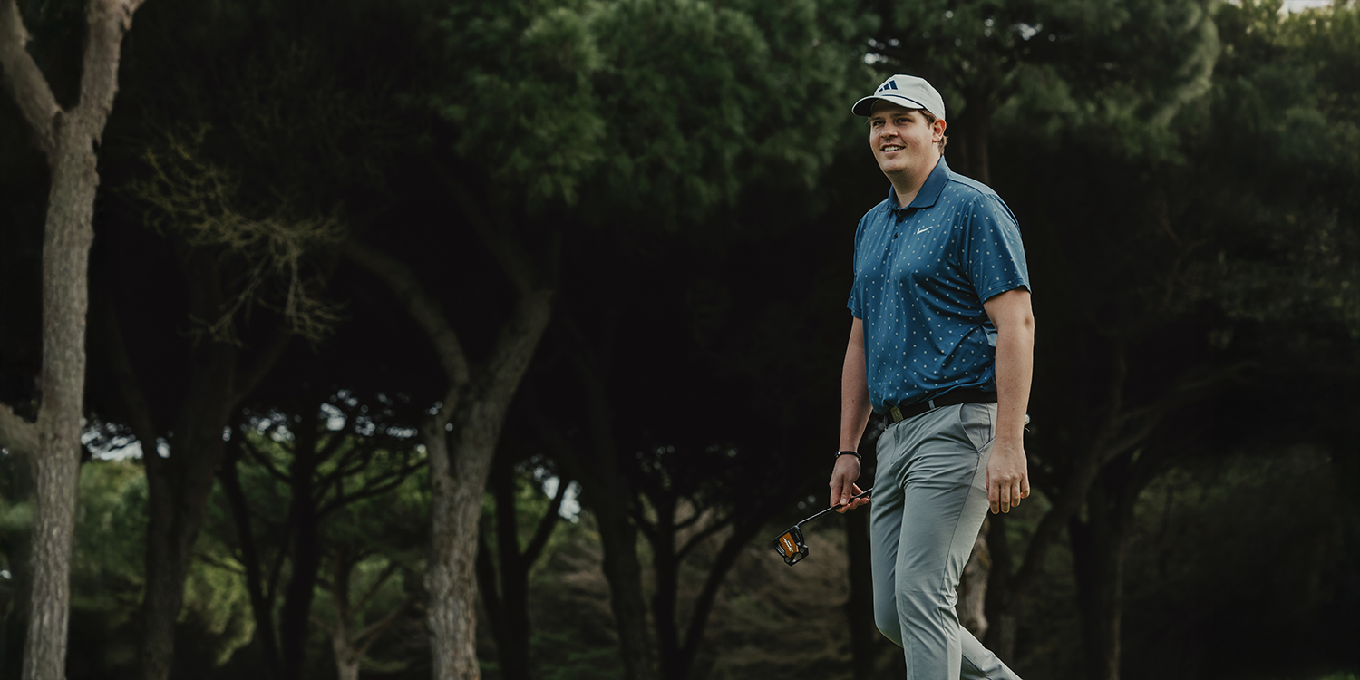 A person is walking on a golf course with a golf club in hand, dressed in a blue polo shirt, grey trousers, and a cap, surrounded by trees.