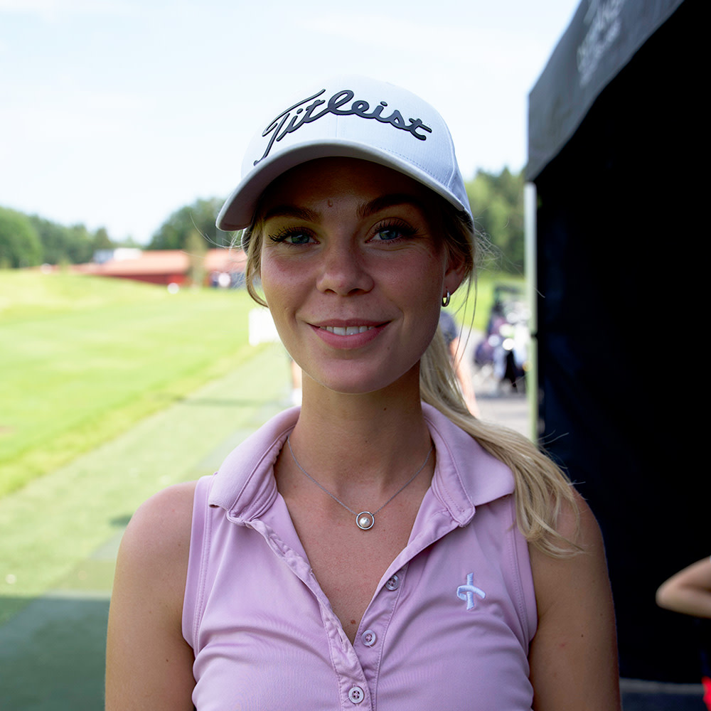 Kvinna med ljus keps med texten "Titleist" och ljusrosa pikétröja står utomhus vid en golfbana och ler mot kameran.