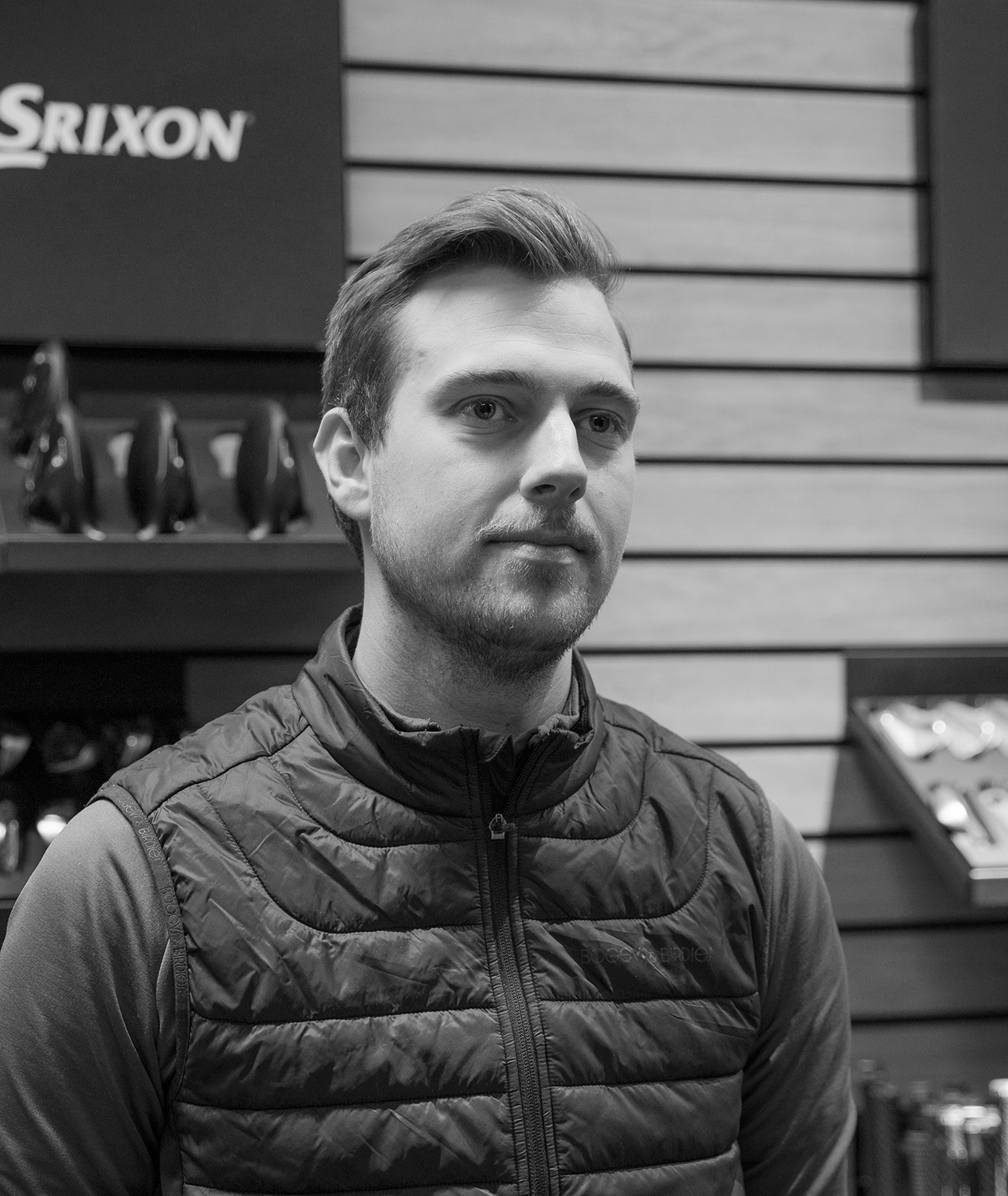 Man med kort hår och skägg bär en vadderad jacka, står inomhus framför en vägg med Srixon-logotyp och golfklubbor i bakgrunden.