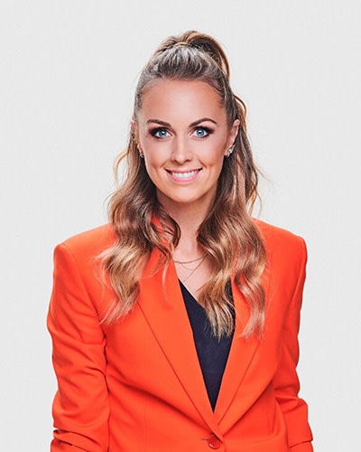 Kvinna med långt, vågigt hår och hästsvans ler och bär orange kavaj över svart topp mot ljus bakgrund.