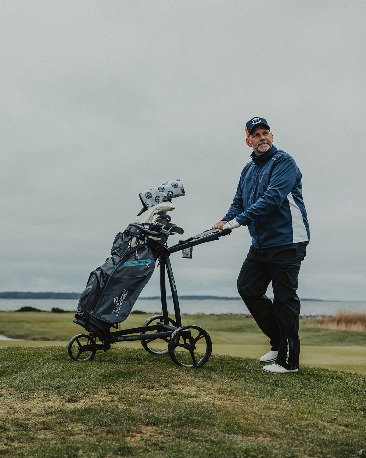 Man i blå jacka och keps skjuter en golfvagn med golfklubbor på en grön golfbana nära vattnet under molnig himmel.