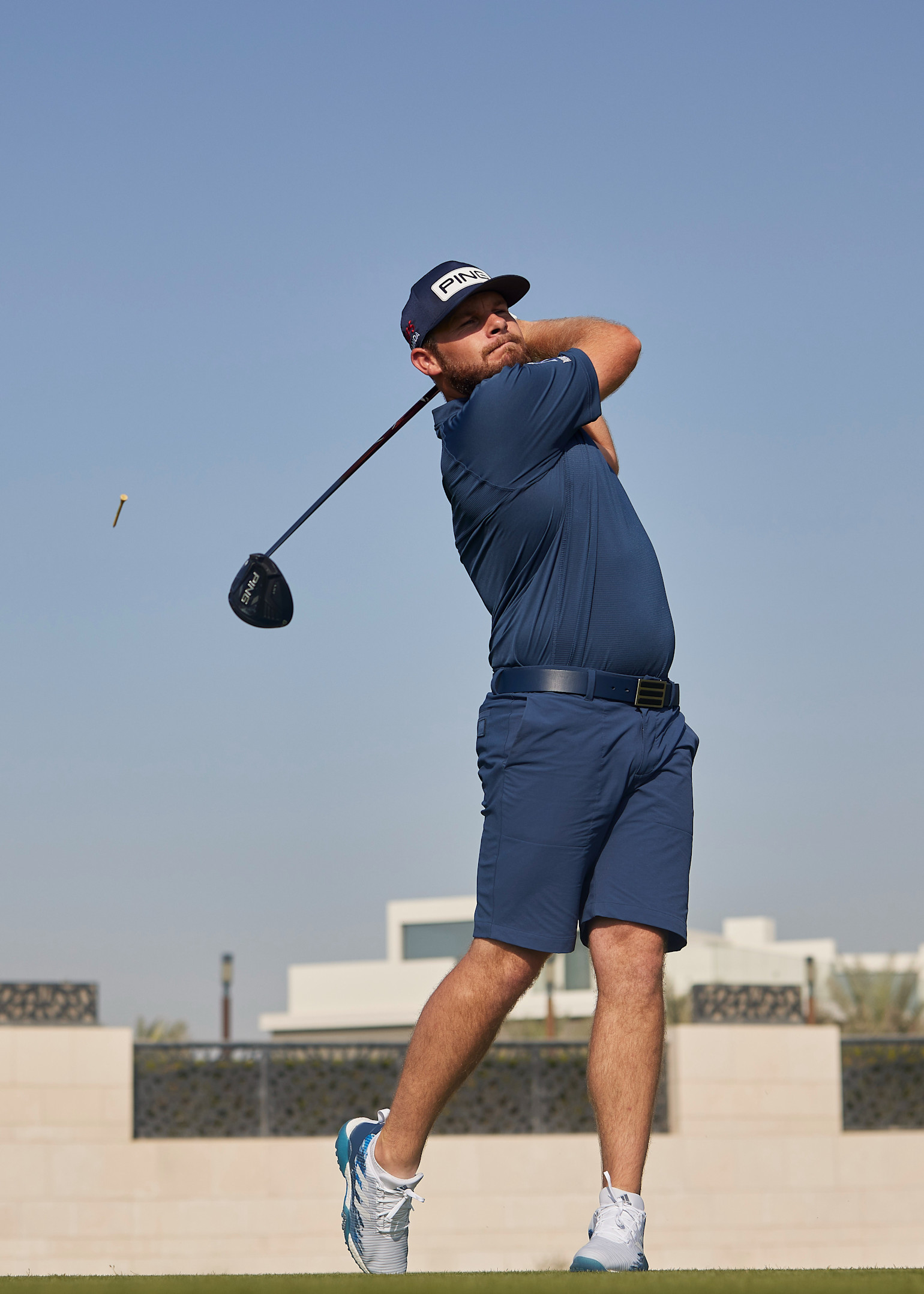 Man i blå golfkläder svingar en golfklubba utomhus på en golfbana med blå himmel i bakgrunden.
