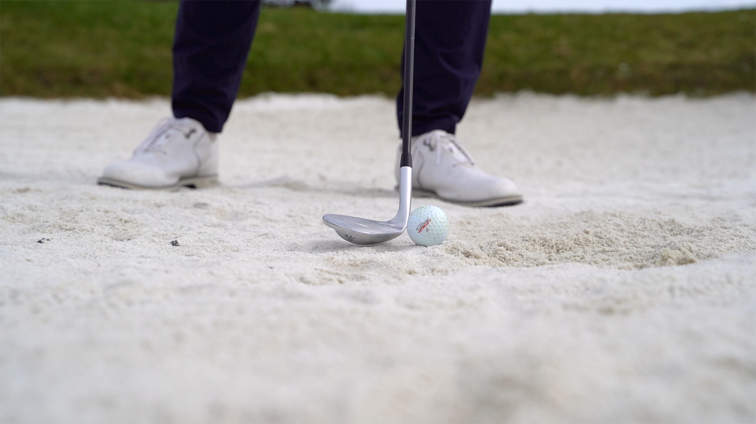 Golfklubba riktas mot en golfboll i en sandbunker, med en person i vita skor och mörka byxor i bakgrunden.