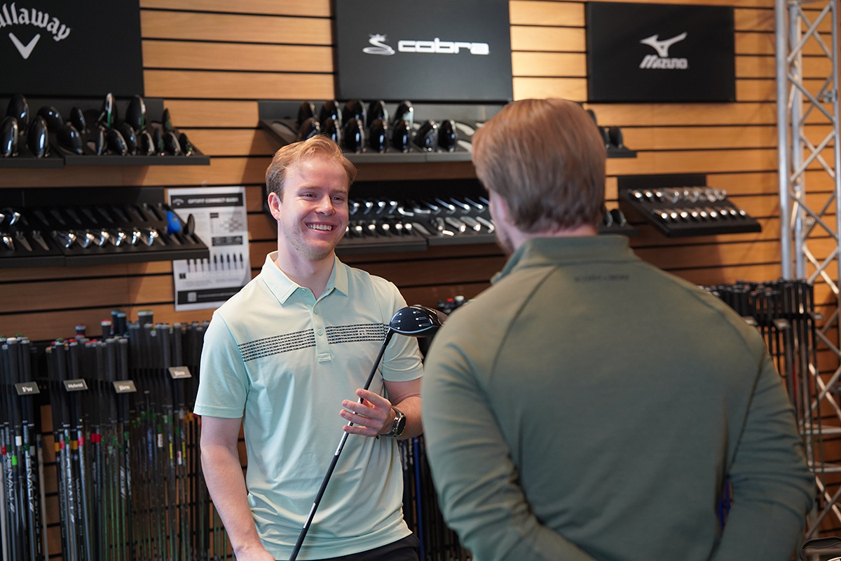 Två män samtalar i en golfbutik, en håller i en golfklubba, omgivna av golfklubbor och märkeslogotyper på väggen.