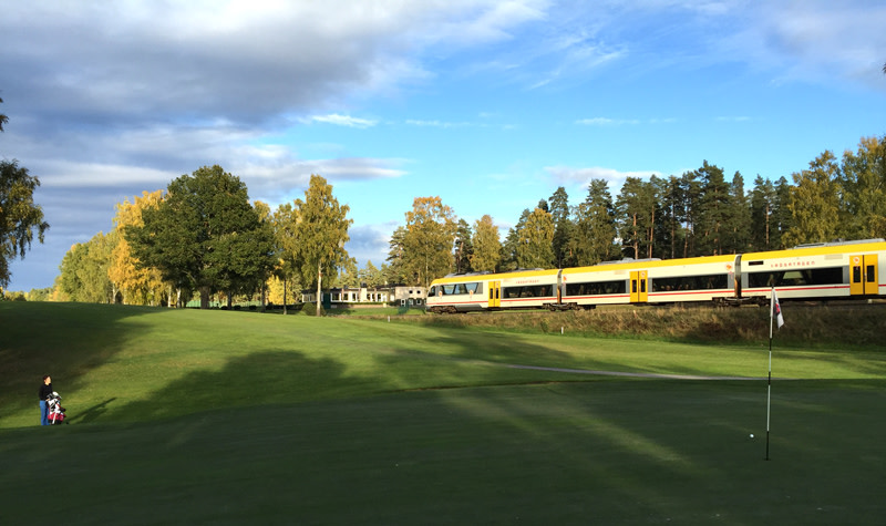 Ett gult och vitt tåg passerar genom ett grönt golfområde med en golfspelare och flagga i förgrunden.