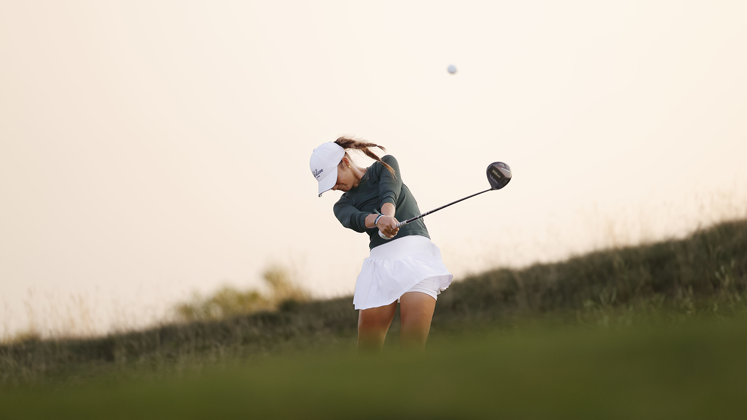 Kvinna i vit keps och kjol svingar en golfklubba och slår en golfboll på en grön golfbana i solnedgång.