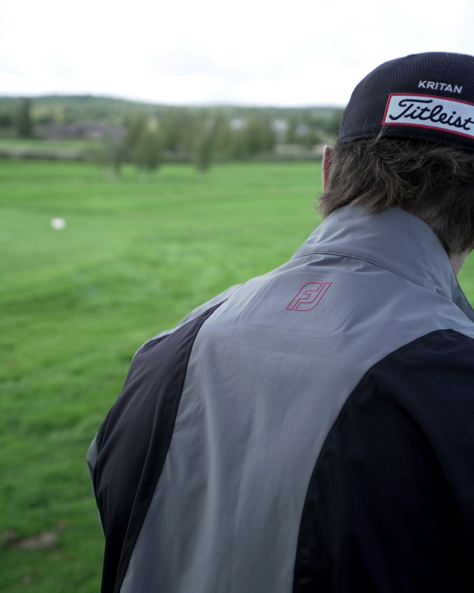 Person med grå och svart jacka och svart Titleist-keps står på en golfbana med grönt gräs i bakgrunden.