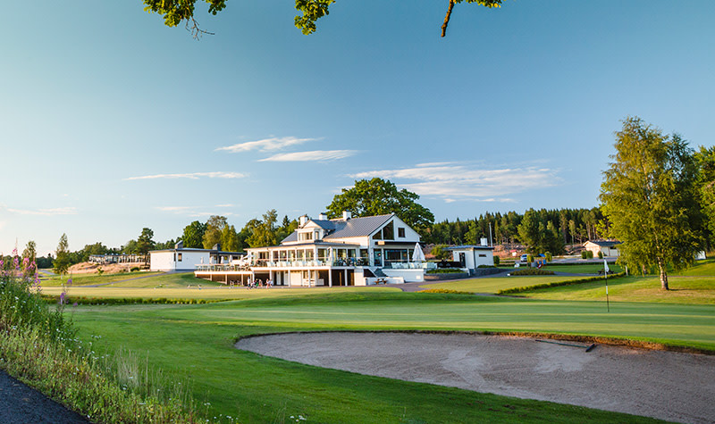 Vit klubbhus med stora fönster och terrass står vid en grön golfbana med sandbunker, omgiven av träd under klar himmel.
