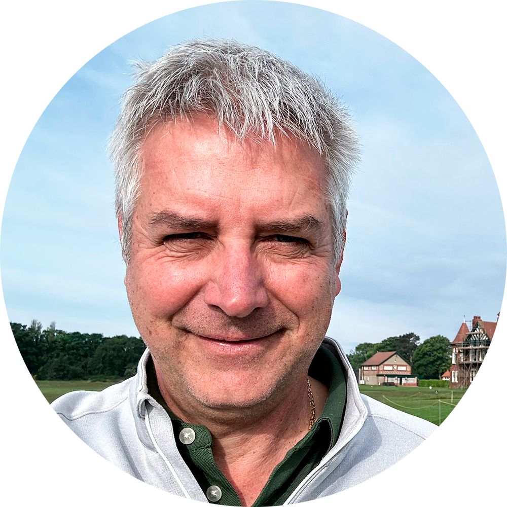 Leende man med grått hår står utomhus framför gräsmatta och hus under en ljusblå himmel.