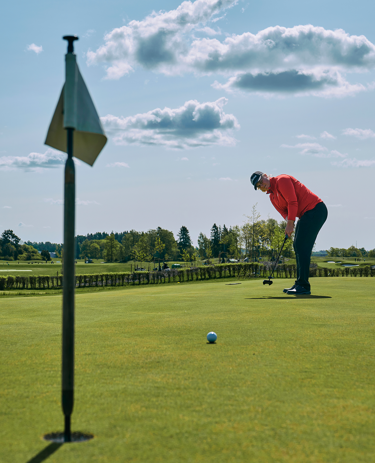 Golfspelare i röd tröja puttar en boll mot hålet på en grön golfbana under en blå himmel med moln.