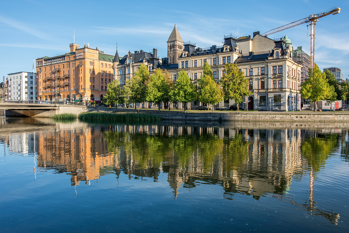 Flera äldre byggnader och träd speglas i en lugn kanal under en klarblå himmel, med en byggkran i bakgrunden.