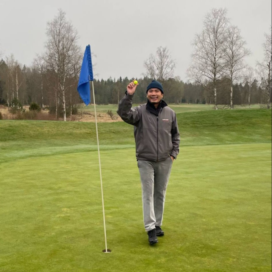 Man ler och håller upp en golfboll bredvid ett hål med blå flagga på en golfbana, omgiven av träd och gräs.