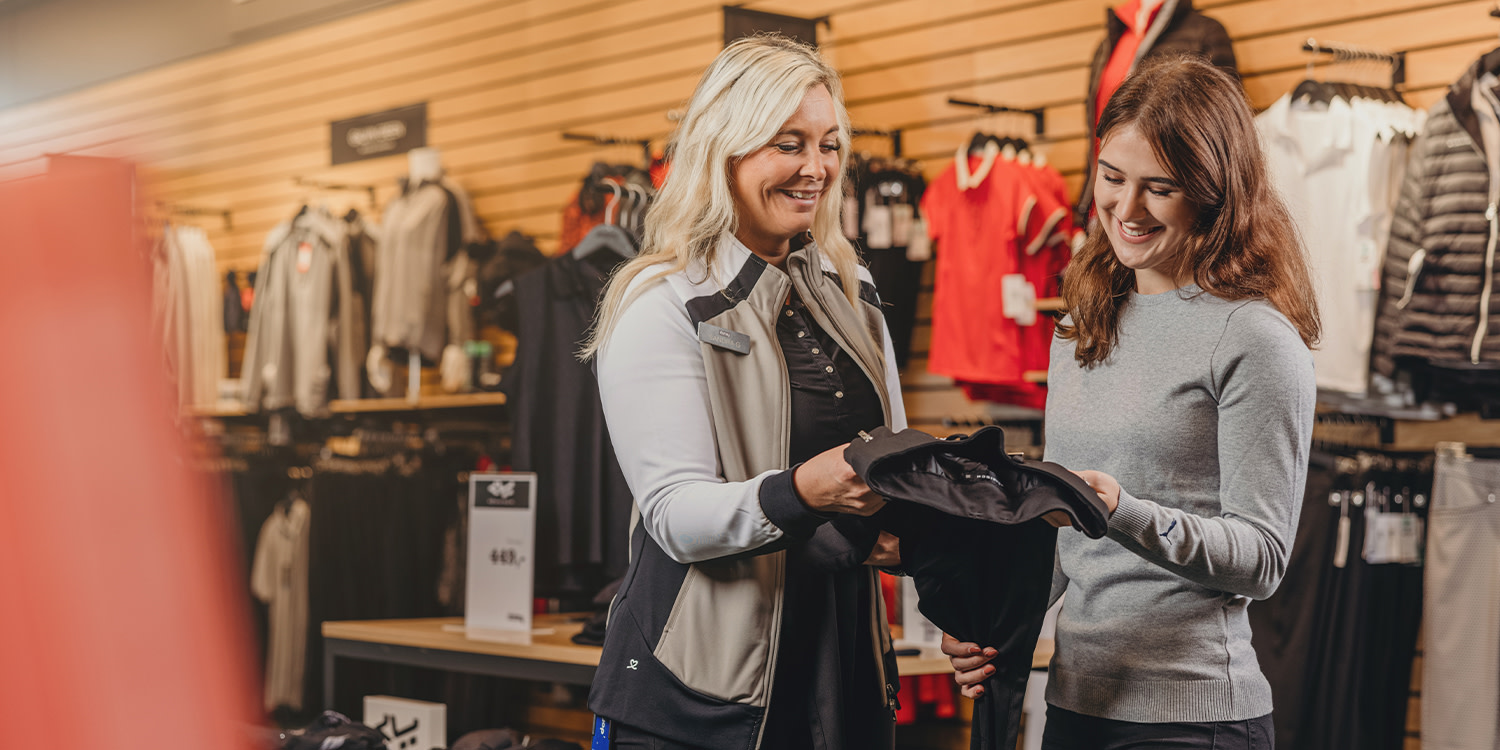 Two women are looking at and smiling at a piece of clothing in a sports shop, surrounded by clothes on hangers and shelves.