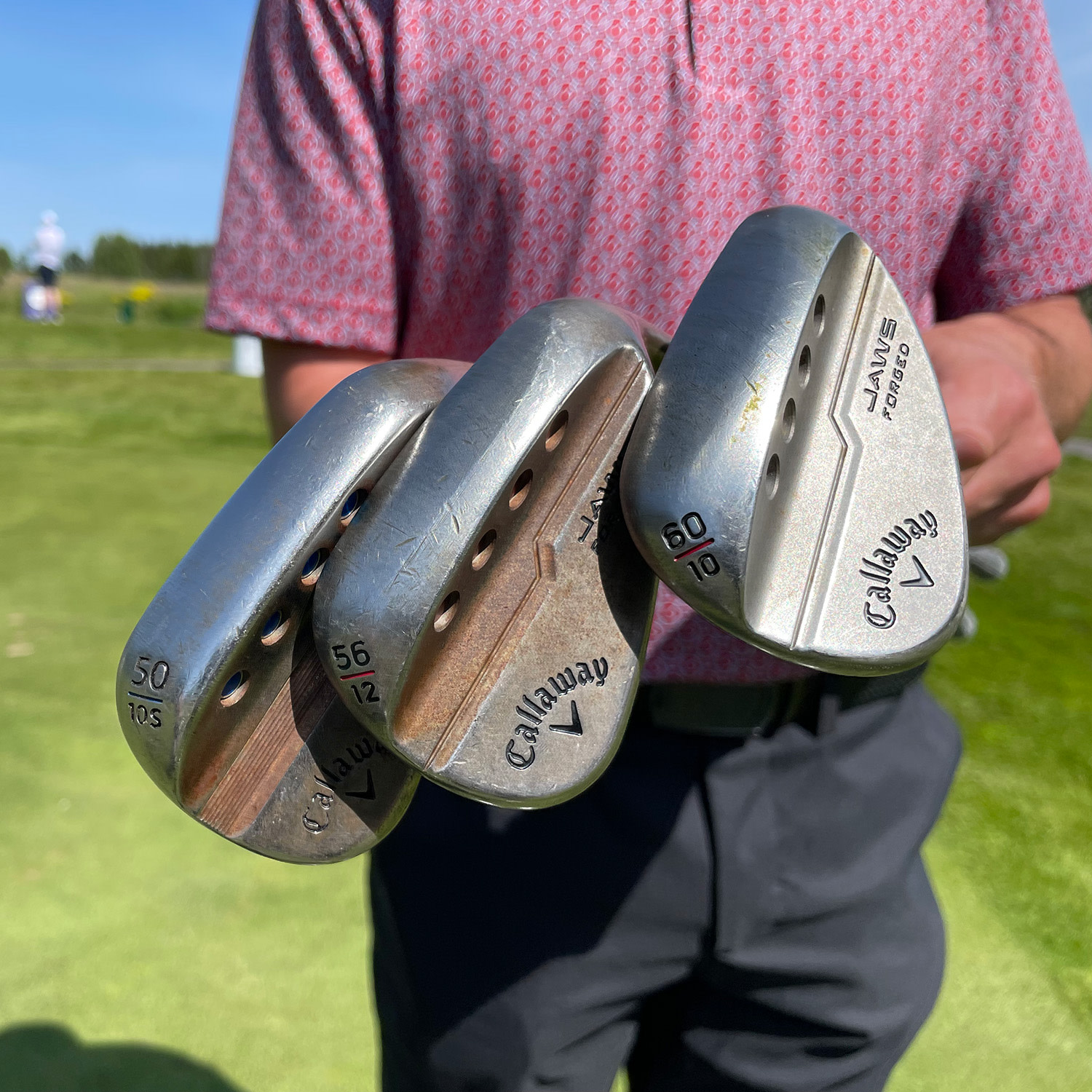 En person håller tre Callaway golfklubbor med olika loftgrader, stående på en golfbana i solsken.