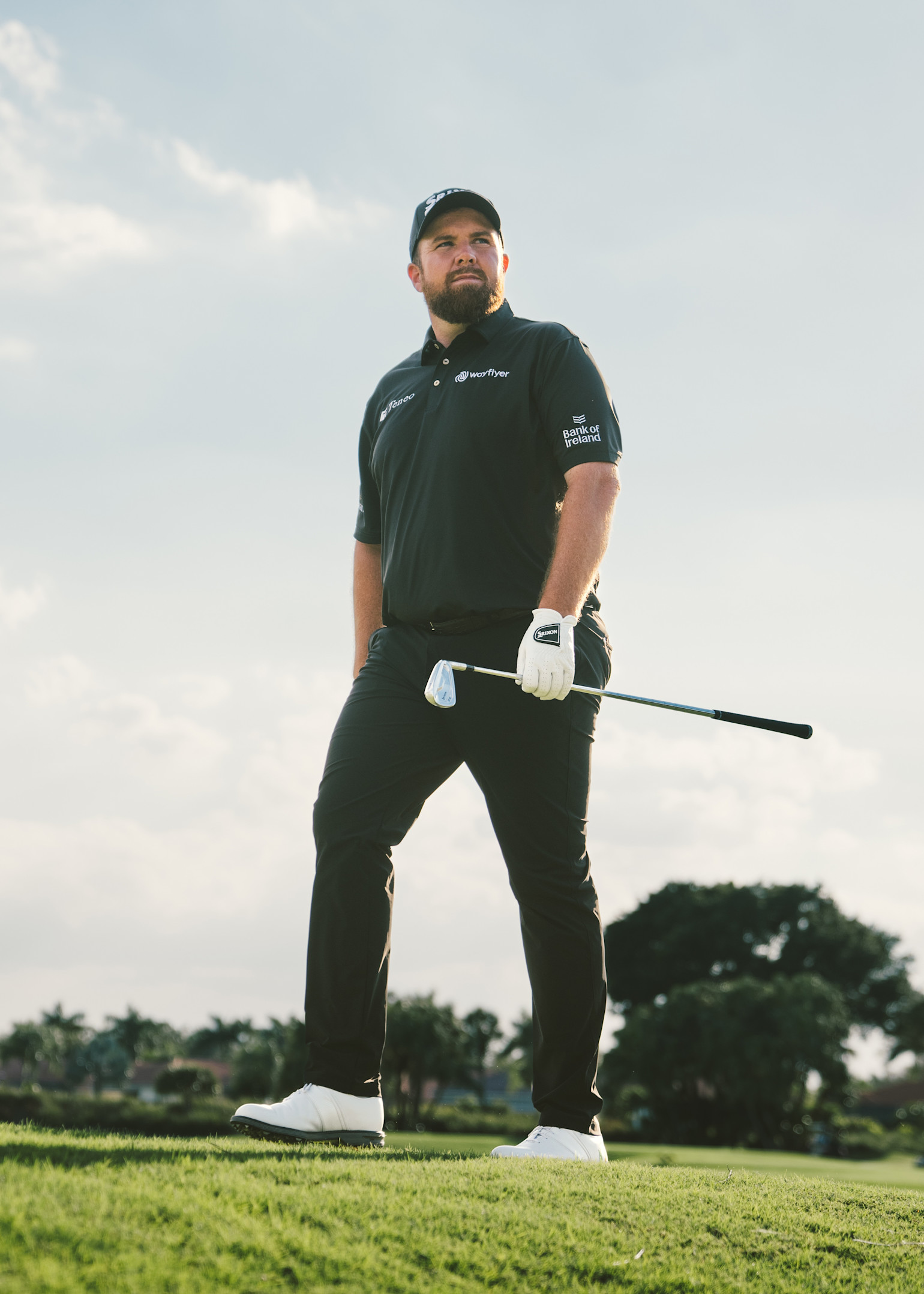 En man i svart golfkläder och keps står på en golfbana med en golfklubba i handen, träd i bakgrunden.