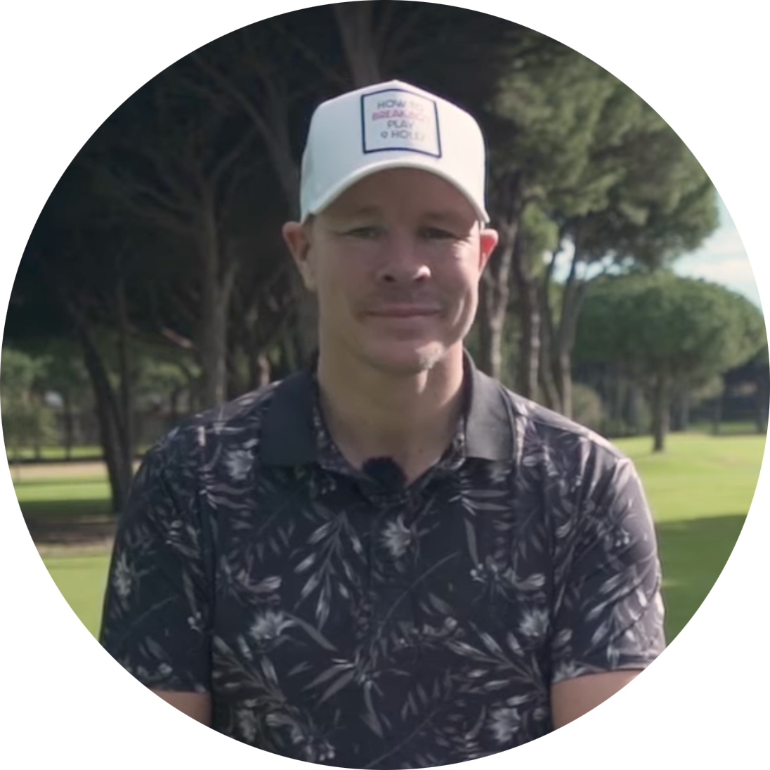 Man med vit keps och mönstrad pikétröja står utomhus på en golfbana med träd i bakgrunden.