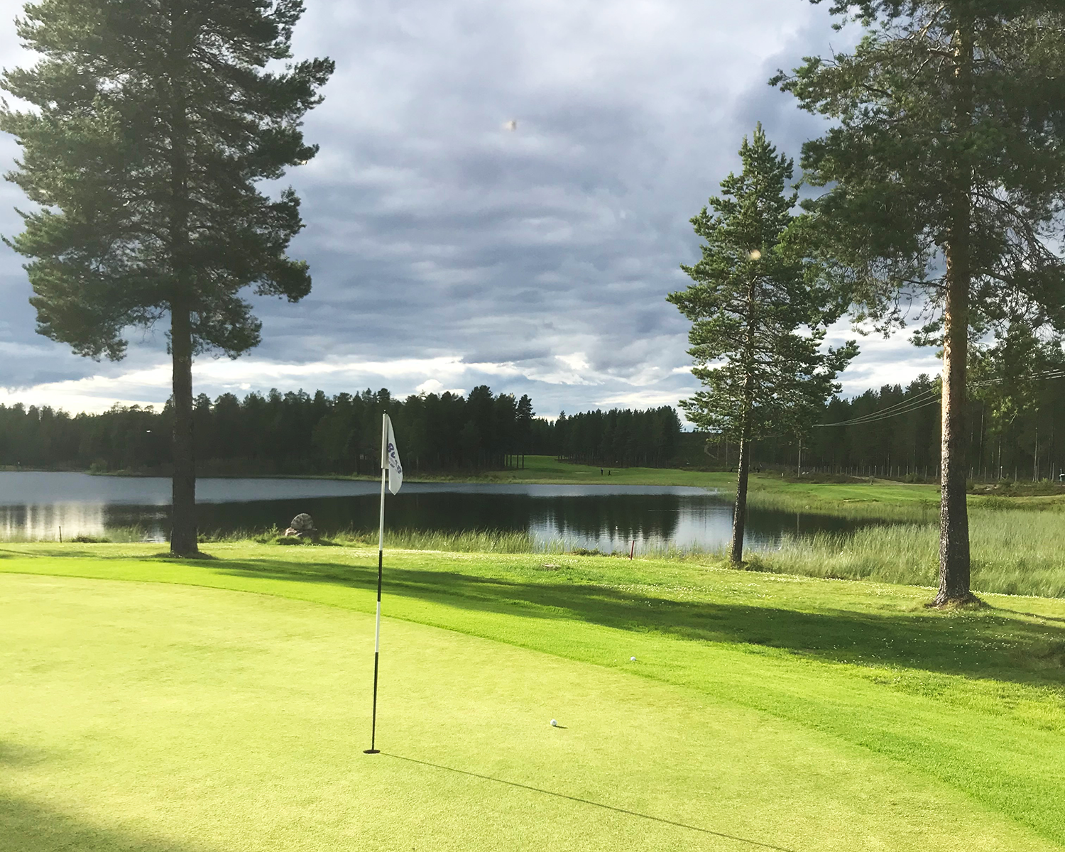 Golfhål med flagga på green, omgiven av träd och sjö under molnig himmel.