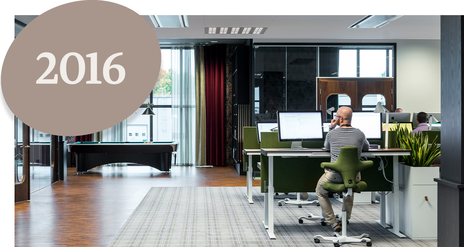 A person is sitting at a desk working on a computer in a modern open-plan office. There is a pool table in the background, and the text "2016" is visible.