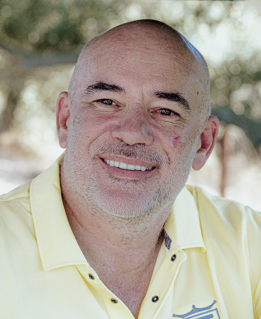 Skallig man med grått skägg ler och bär en gul pikétröja, suddig grön natur i bakgrunden.