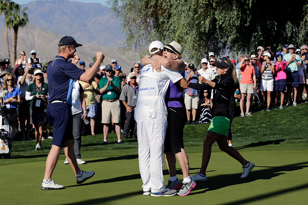 Två personer kramas på en golfbana medan andra firar och en publik applåderar i bakgrunden under soligt väder.
