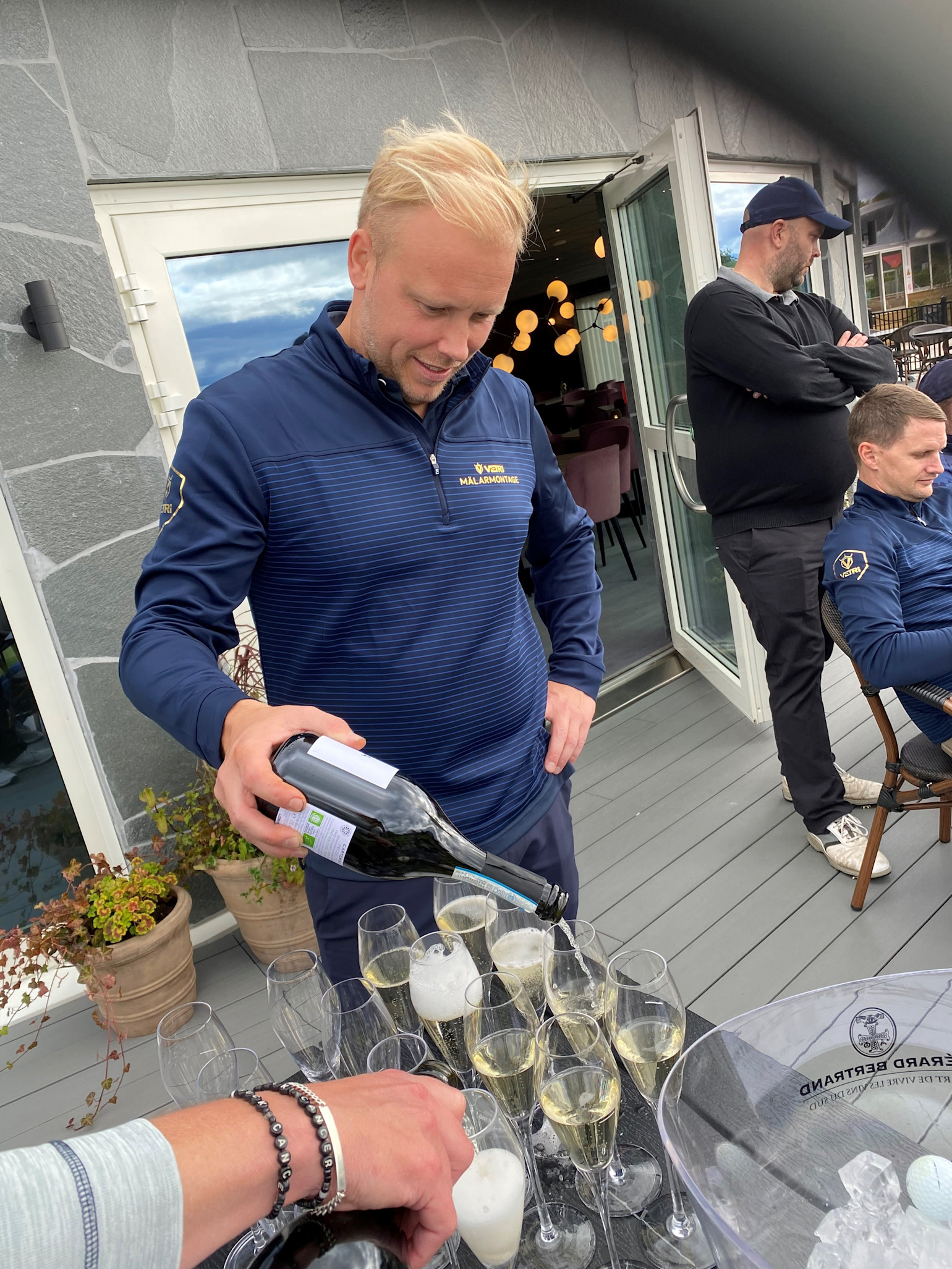 Man i blå tröja häller mousserande vin i glas utomhus på en terrass, med fler personer och öppna dörrar i bakgrunden.